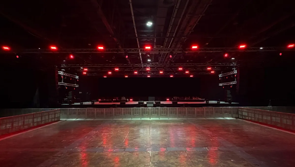 Concert stage set up in an empty hall before doors, with LED screens, lighting trusses and a crowd barrier in front of the stage.